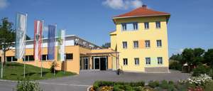 Ein gelbes Gebäude mit Fahnen davor, eine Einfahrt mit einem Garten davor und ein klarer blauer Himmel.