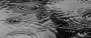 Schwarz-Weiß-Bild von Wassertropfen, die auf eine Oberfläche spritzen und Wellen und Wirbel erzeugen. Die Tropfen variieren in Größe und Intensität, einige überlappen sich.