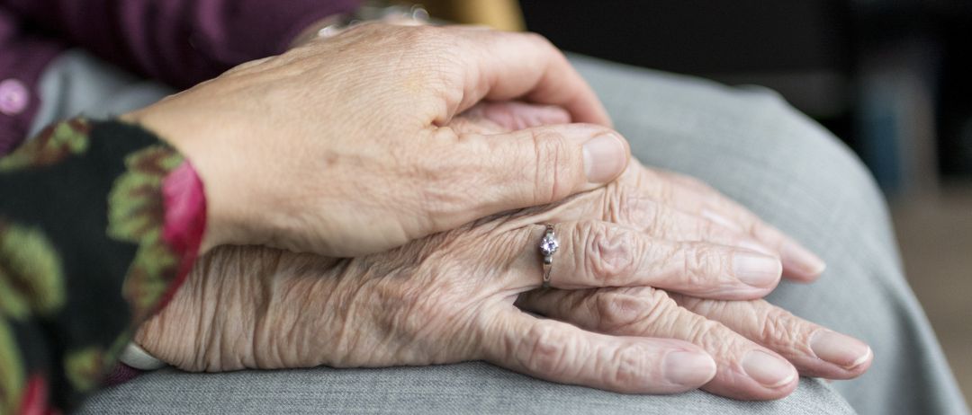 An elderly person wearing a ring holds hands with another person, both hands are wrinkled and the fingers are slightly curled.