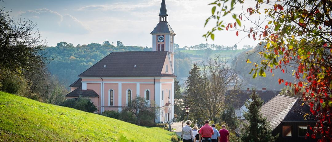 Eine Gruppe von Menschen geht einen Hügel hinauf zu einer Kirche mit Glockenturm. Die Kirche hat eine rosa Fassade und ist von Bäumen umgeben. In der Ferne gibt es Hügel und einen klaren Himmel.