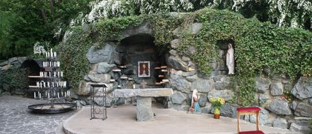A small stone chapel with a statue of Mary, surrounded by greenery, has a stone altar, a candle stand, and a small wooden stool with a red cushion.