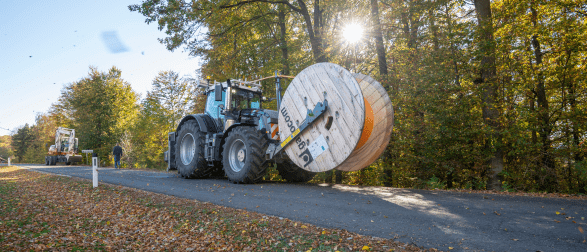 Ein großer Traktor mit zwei Drahtspulen fährt auf einer Straße mit Herbstbäumen auf beiden Seiten. Eine Person geht in der Nähe vorbei.