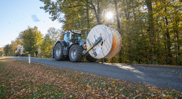 Ein großer blauer Traktor mit einer Spule aus orangefarbenem und weißem Kabel steht auf einer von Bäumen gesäumten Straße. Eine Person geht am Straßenrand.