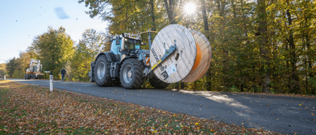 Ein großer blauer Traktor mit einer Spule aus orangefarbenem und weißem Kabel steht auf einer von Bäumen gesäumten Straße. Eine Person geht am Straßenrand.