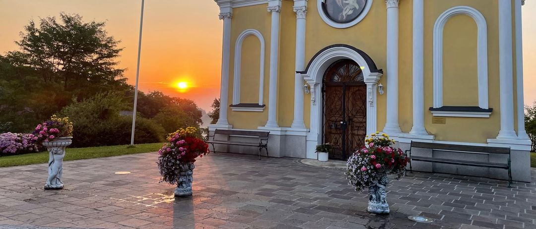 Eine gelbe Kirche mit einem Turm, verziert mit weißen Umrandungen und Bögen, geschmückt mit Blumenvasen und einer Bank draußen. Die Sonne geht hinter den Bäumen in der Ferne unter.