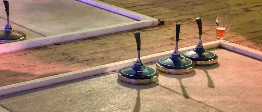 Drei blaue Curlingsteine mit silbernen Griffen sind auf einer glatten Fläche in einer Reihe aufgestellt, wobei Schatten auf der Oberfläche geworfen werden.