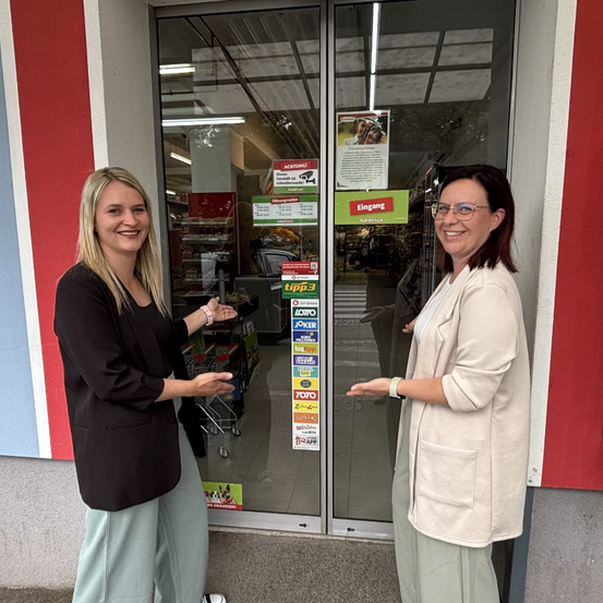 Zwei Frauen stehen lächelnd vor dem Eingang eines Geschäfts, eine hält ein Schild mit der Aufschrift 'tipp3'. Hinter ihnen zeigen die Glastüren des Geschäfts verschiedene Werbeplakate.