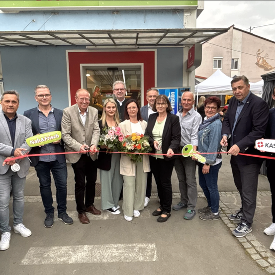 Eine Gruppe von Menschen steht vor einem Geschäft, hält ein Band und Blumen und feiert wahrscheinlich eine Eröffnung.