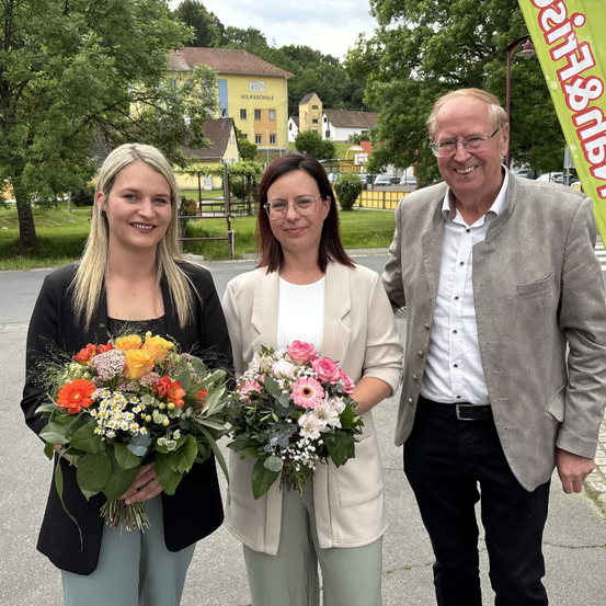 Drei Menschen stehen an einem sonnigen Tag zusammen und lächeln. Zwei Frauen halten Blumensträuße. Ein Mann steht zwischen ihnen. Sie sind draußen in der Nähe von Gebäuden und Bäumen.