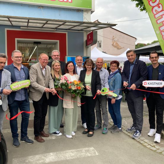Eine Gruppe von Menschen steht vor einem Geschäft, lächelt und hält eine Schleife für eine Eröffnungsfeier. Einige tragen Brillen, andere haben Blumen. Das Geschäft hat ein Schild mit grünem Text.
