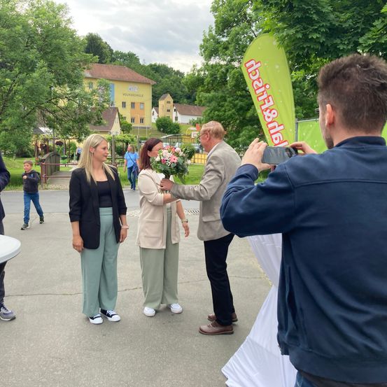 Zwei Frauen und ein Mann stehen vor einem Mann, der ein Foto mit einer Kamera macht. Die Frau hält einen Blumenstrauß. Dahinter steht ein Banner mit dem Text "Freizeit & Spaß". In der Ferne sind Bäume, Häuser und eine Brücke zu sehen.