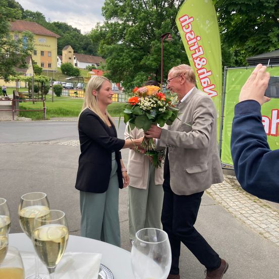 Eine Outdoor-Veranstaltung, eine Frau in einem schwarzen Blazer übergibt einem Mann im grauen Anzug Blumen. Dahinter stehen zwei Frauen mit einem Hintergrund. Im Hintergrund befinden sich Häuser, Bäume und eine Person, die ein Foto macht.