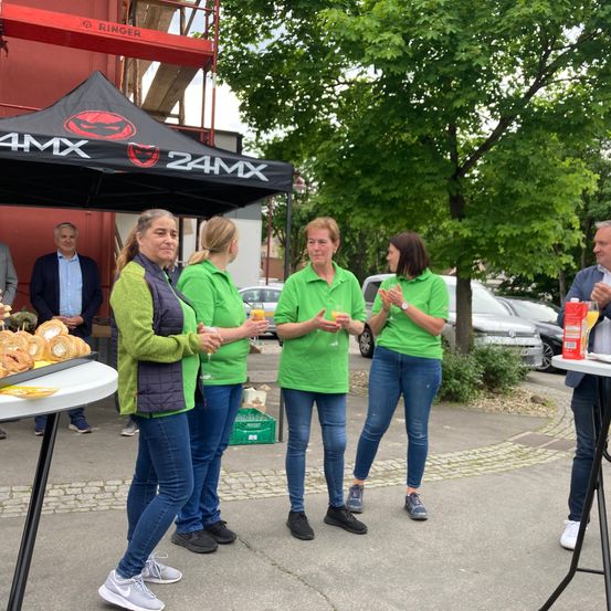 Fünf Frauen in grünen Shirts stehen neben einem Tisch und halten Getränke. Sie scheinen zu klatschen oder zu jubeln. Ein schwarzes Zelt mit dem 24MX-Logo steht hinter ihnen. Bäume, geparkte Autos und ein Gebäude sind im Hintergrund.