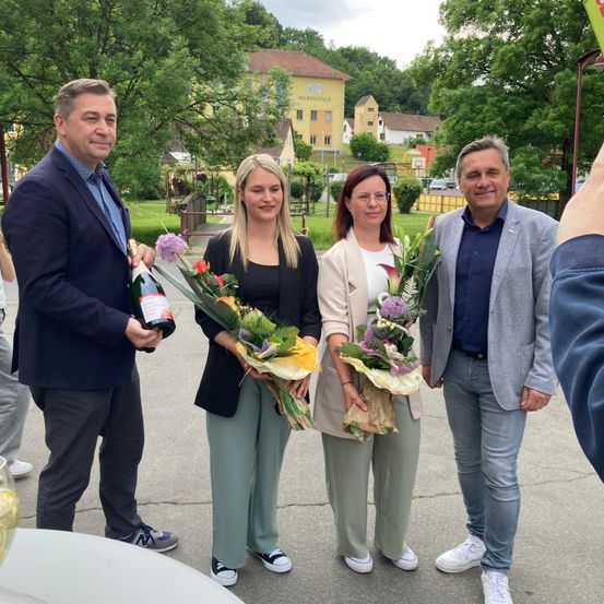 Vier Erwachsene stehen in einem Park, halten Blumen und eine Flasche. Der Mann auf der linken Seite hält eine Flasche Champagner. Sie stehen auf einer Betonfläche. Bäume und Gebäude sind im Hintergrund.