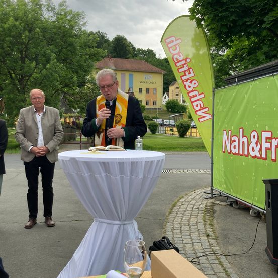 Ein Priester hält eine Rede bei einer Veranstaltung im Freien. Hinter ihm steht ein Mann in der Nähe eines Tisches mit einem Buch und einem Glas Wasser. Ein Banner mit den Worten 'Nah&Eirich' ist rechts angebracht. Bäume und Gebäude sind im Hintergrund zu sehen.