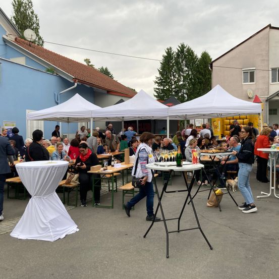 Eine Outdoor-Versammlung mit weißen Zelten und Tischen, an der viele Menschen plaudern, trinken und essen. Ein blaues Gebäude und ein gelbes Zelt im Hintergrund.