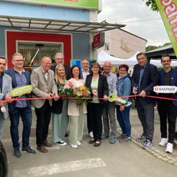 Eine Gruppe von Menschen steht vor einem Gebäude mit einem roten Band. Sie halten Blumen und einen Bandschneider. Sie lächeln und posieren für ein Foto.