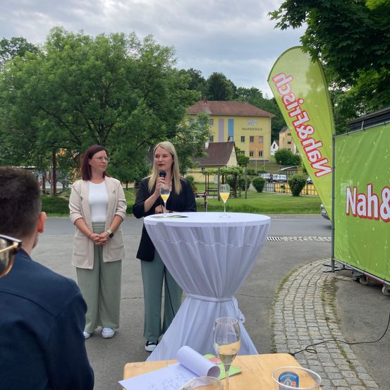Zwei Frauen sprechen bei einer Veranstaltung, mit Weingläsern auf einem Tisch. Dahinter steht ein Gebäude mit gelber Fassade und grünem Schild. Der Ort ist im Freien mit Bäumen und einem Weg.