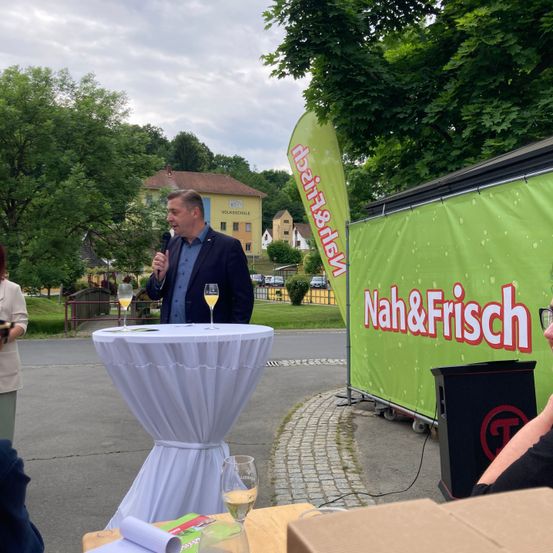 Ein Mann steht an einem Rednerpult mit einem Mikrofon und spricht vor einem grünen Banner, das 'Nah&Frisch' besagt. Zwei Weingläser stehen auf dem Podium. Dahinter befindet sich ein Tisch mit einer weißen Tischdecke, auf dem zwei weitere Weingläser stehen. Im Hintergrund sind Bäume, Gebäude und eine Straße zu sehen.