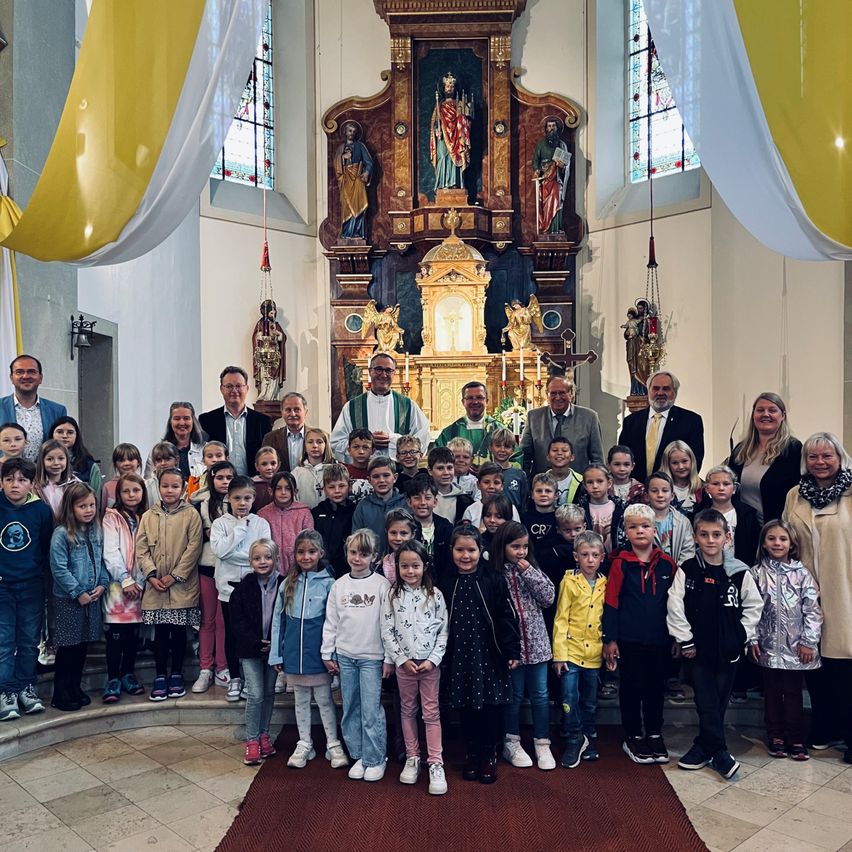 Eine Gruppe von Kindern und Erwachsenen steht vor einem Altar in einer Kirche, mit Statuen und einem Priester im Hintergrund sichtbar.