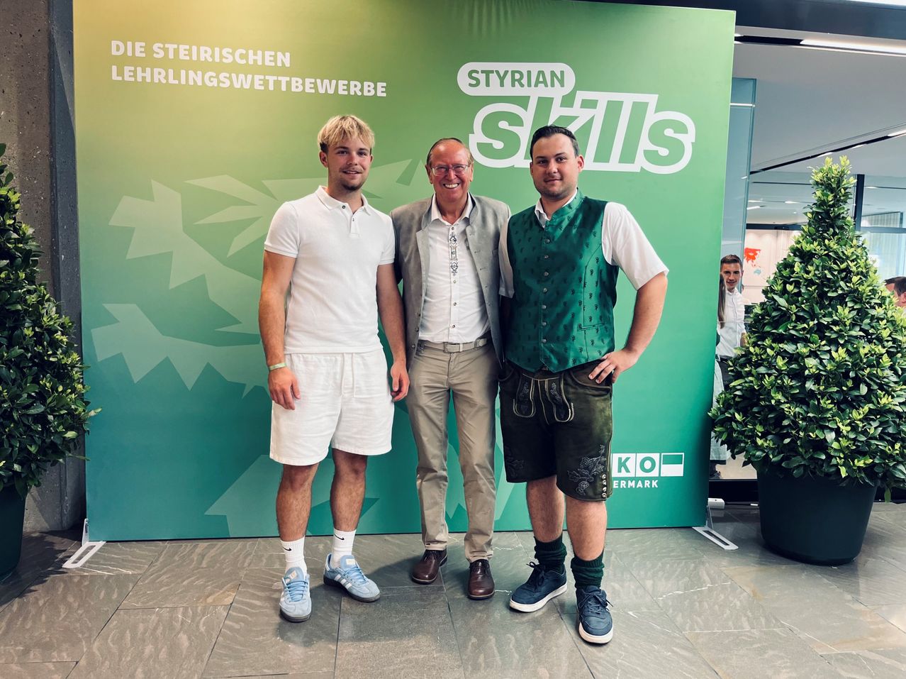 Drei Männer posieren für ein Foto vor einem grünen Banner. Der Mann auf der linken Seite trägt Weiß, der Mann in der Mitte Grau und der Mann auf der rechten Seite Grün.