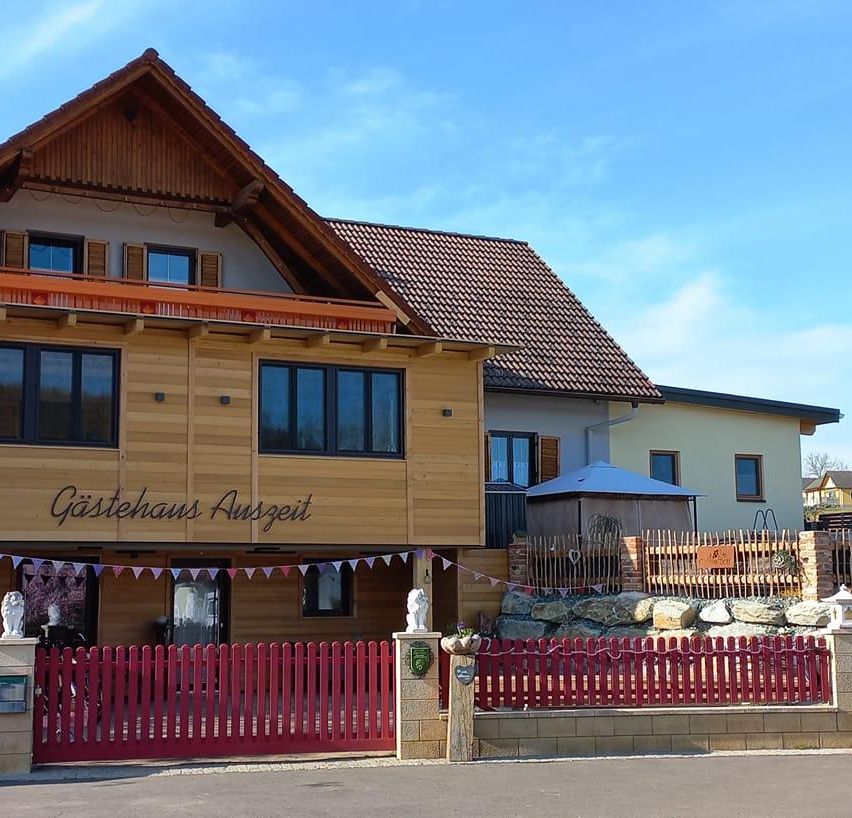 Ein Haus mit braunem Ziegeldach und Holzwänden. Es hat viele Glasfenster und einen Balkon. Es steht Gastehaus Auszeit darauf geschrieben. Vor dem Haus steht ein roter Zaun.