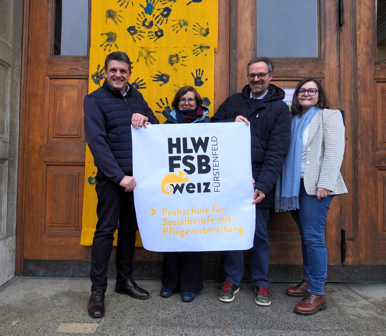 Vier Personen stehen vor einer Holztür mit einem gelben Banner und halten ein weißes Banner mit dem Text 'HLW FSB Weiz Furstenfeld' und dem Slogan 'Fachschule für Sozialberufe'.