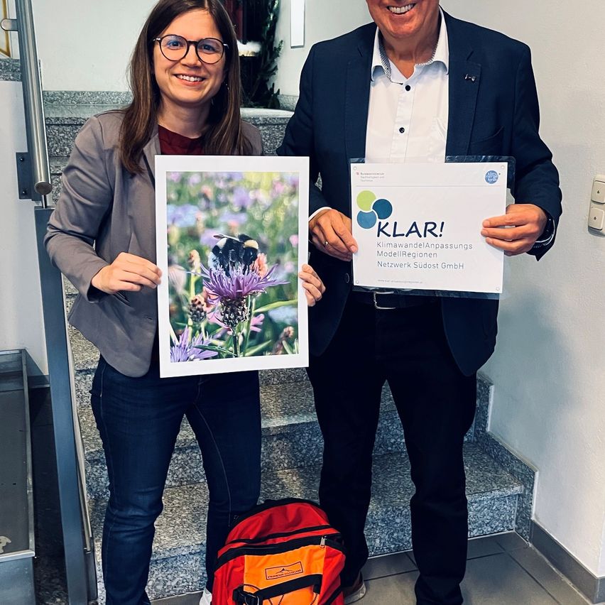 Eine Frau und ein Mann stehen auf einer Treppe mit einem roten Rucksack. Die Frau hält ein gerahmtes Foto eines Hummels auf einer Blume, und der Mann hält ein Zertifikat mit dem Titel 'KLAR!'.