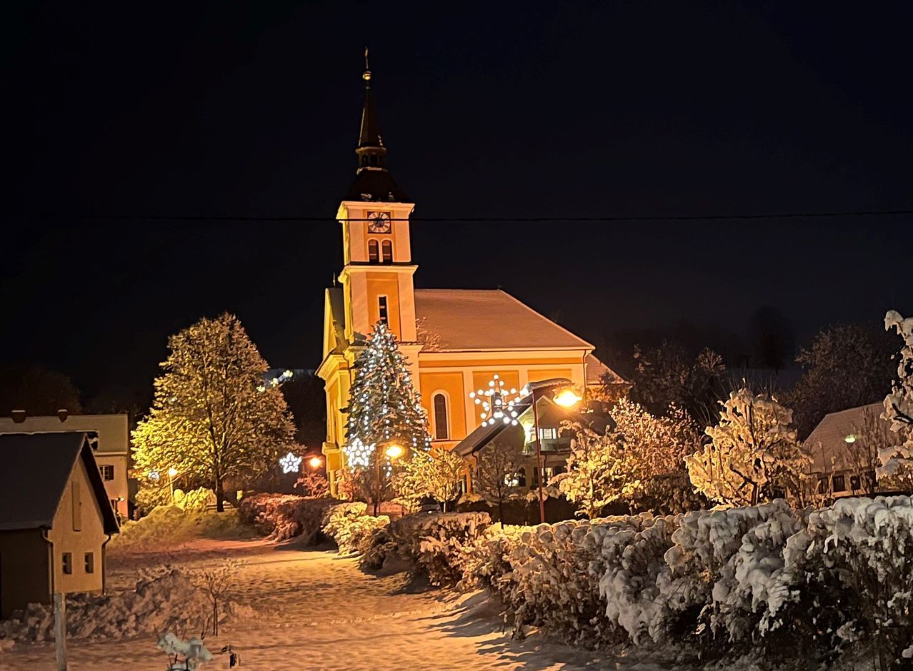 In der Nacht ist eine Kirche mit Glockenturm wunderschön beleuchtet, umgeben von schneebedeckten Bäumen und Pflanzen und einem Weihnachtsbaum davor.