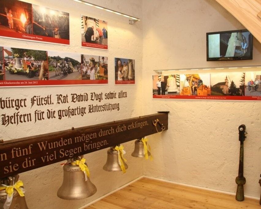 Ein Raum mit Glocken und Bildern an der Wand. Ein deutscher Satz ist auf einem Holzschild über den Glocken geschrieben. Ein Monitor ist an der Wand montiert.