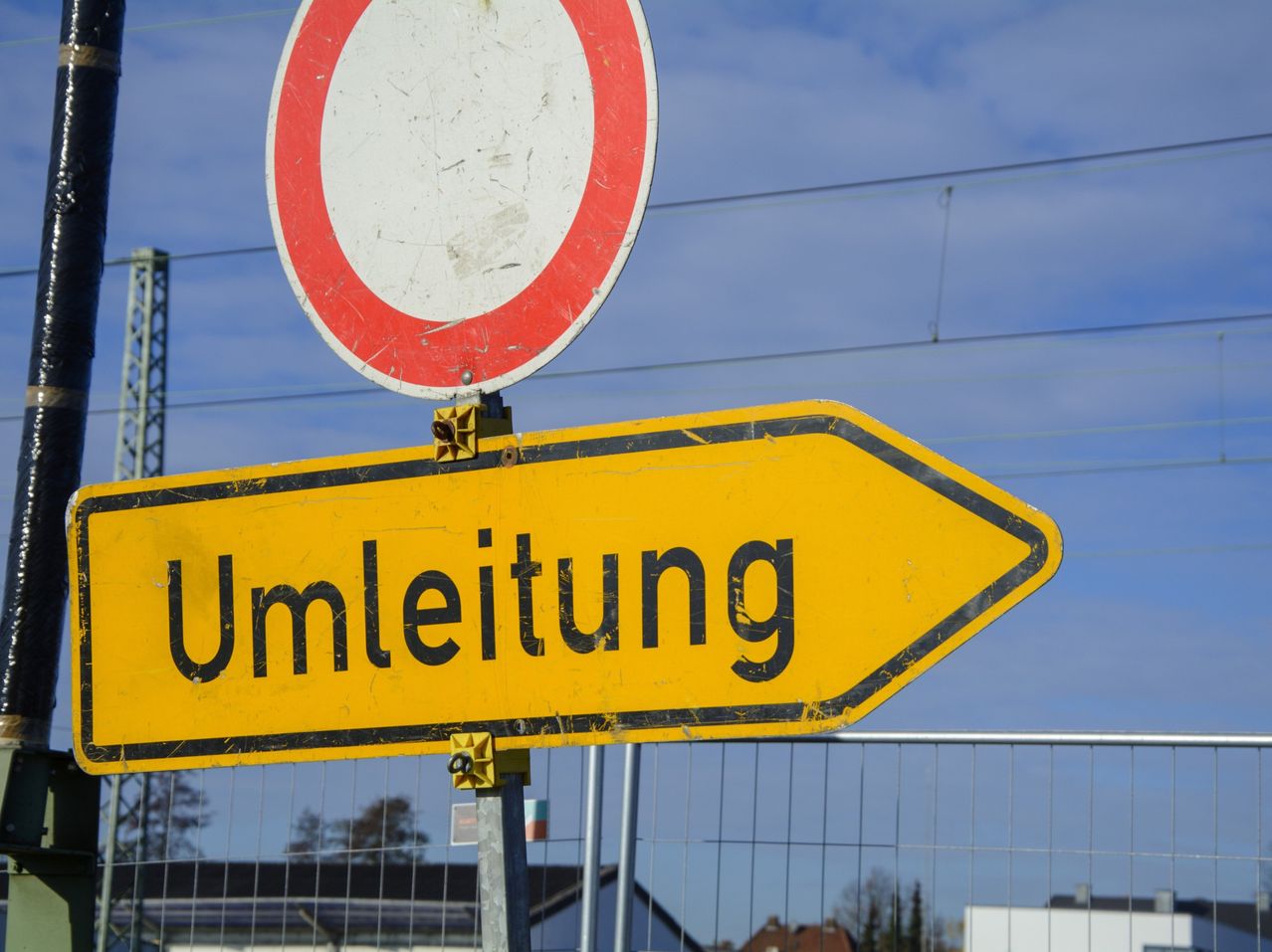 A yellow road sign with the word 'Umleitung' is mounted on a metal pole, indicating a diversion. A round traffic sign is above it. Behind the sign is a fence with a metal wire.