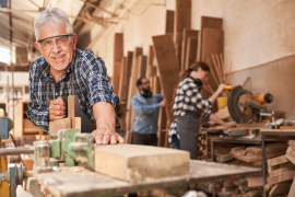 An older man is working on a piece of wood in a workshop. A woman is cutting wood with a circular saw behind him.