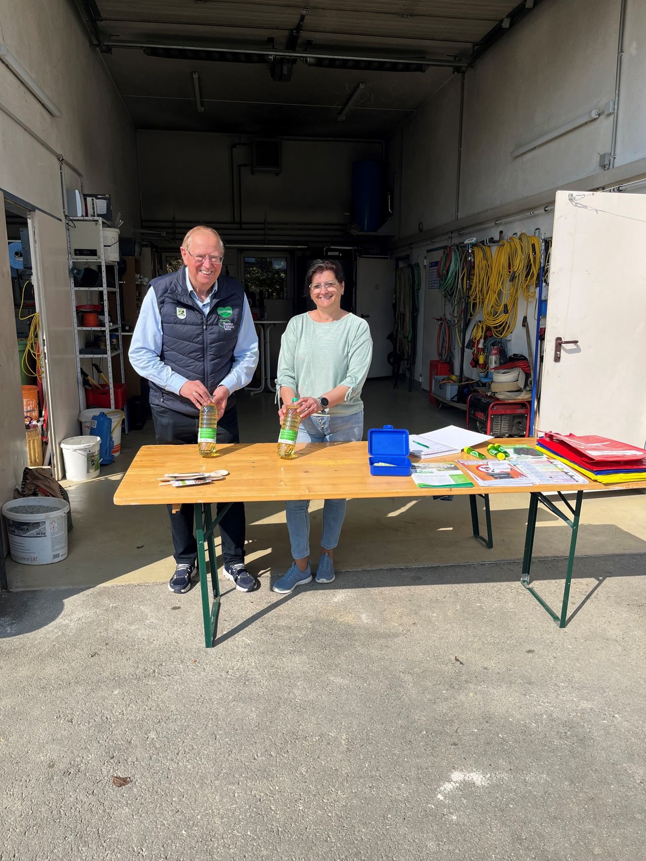 An elderly man and woman stand behind a table in a garage, smiling for a photo. They have two bottles of liquid on the table and papers, a box, and other items.