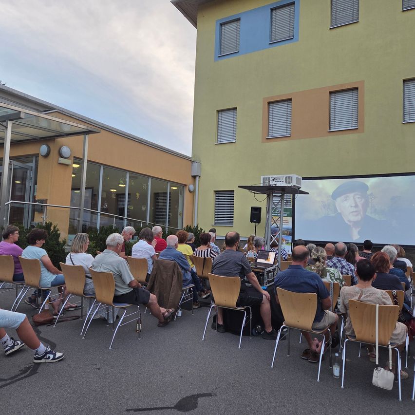 Eine Filmvorführung im Freien, bei der ein Publikum auf Stühlen vor einem Gebäude sitzt und einen Film auf einer Leinwand beobachtet.