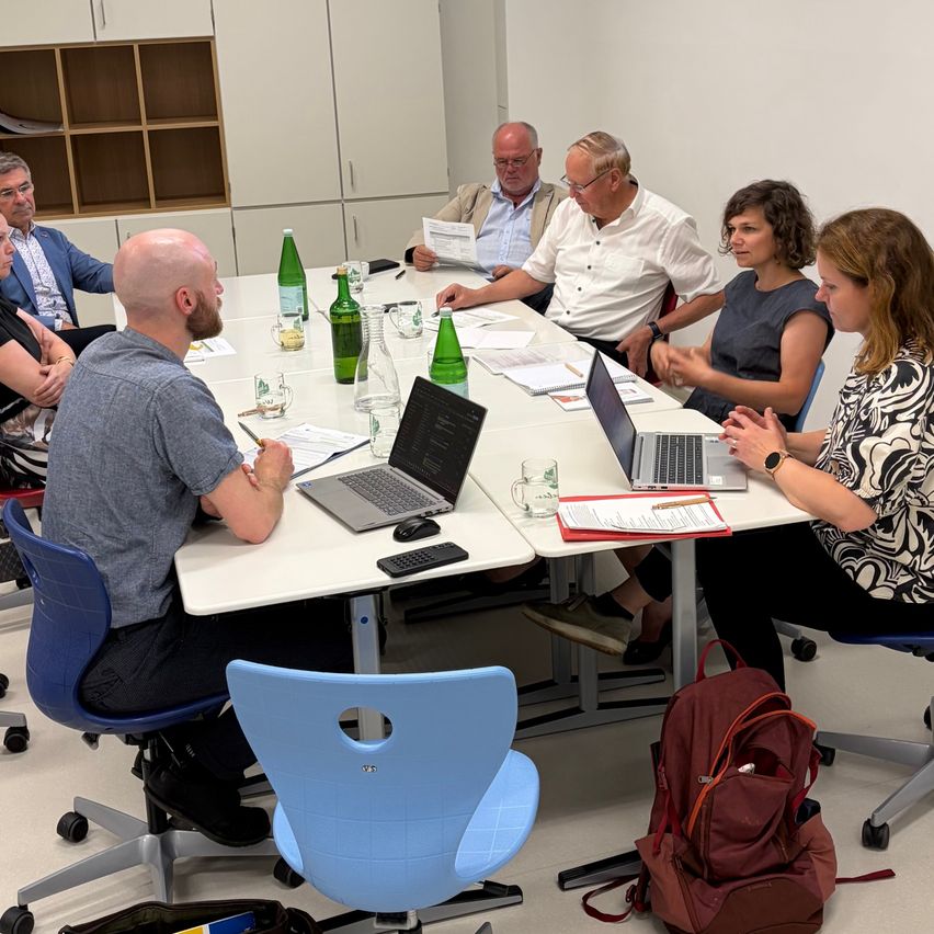 A group of people sit around a table with laptops, papers, and green bottles. They appear to be engaged in a meeting.