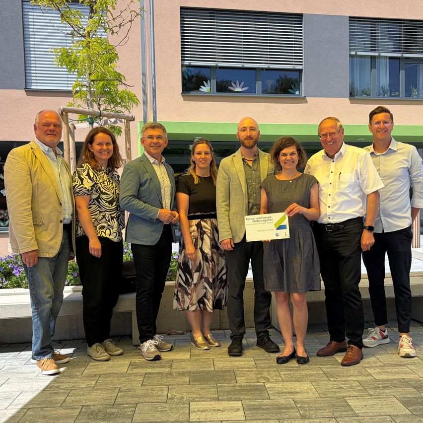 Seven individuals are standing outside a building with one woman holding a certificate. They are all smiling and seem to be posing for a photo.