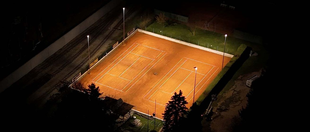 Luftaufnahme eines Tennisplatzes im Freien mit vier Tennisplätzen, beleuchtet von Straßenlaternen bei Nacht.