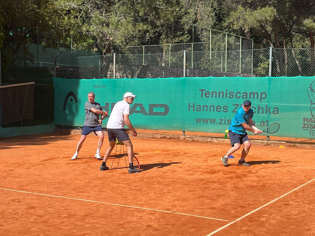 Drei Männer spielen Tennis auf einem Sandplatz, einer schlägt einen Ball und zwei andere beobachten.