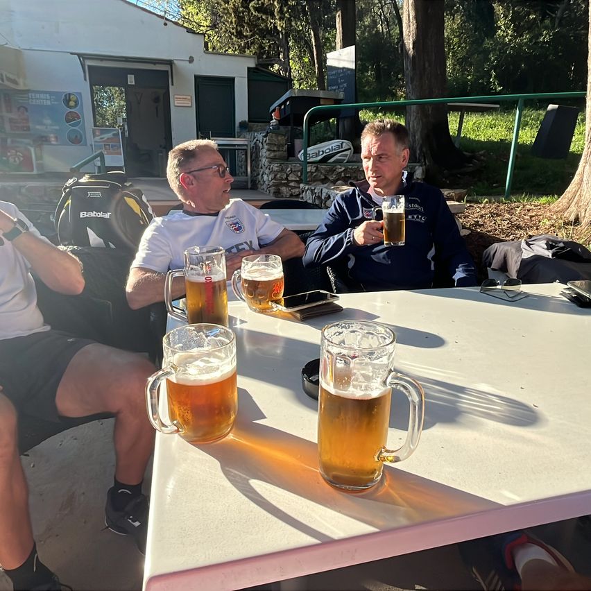 Drei Männer sitzen draußen an einem Tisch, jeder hält ein Glas Bier in der Hand. Einer trägt ein weißes Shirt, der andere eine blaue Jacke und der dritte ein weißes Shirt und schwarze Shorts. Der Tisch hat drei Gläser Bier, und ein Rucksack steht hinter ihnen.