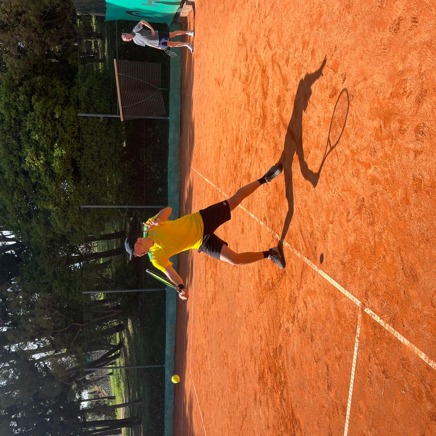 Tennisspieler in gelbem Shirt und schwarzen Shorts spielt Tennis auf einem Sandplatz, in der Luft mit einem Tennisschläger und Schatten auf dem Boden.