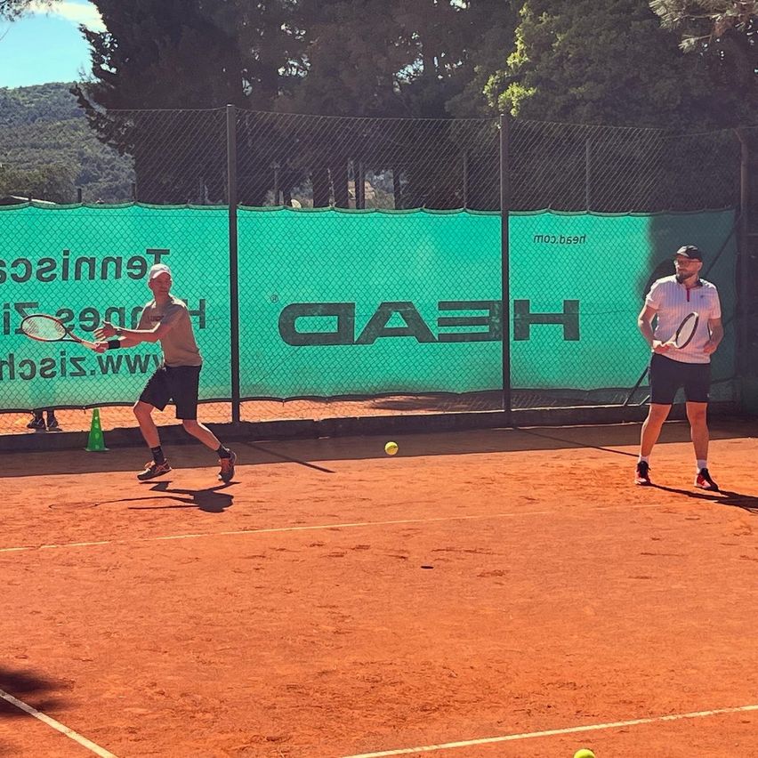 Zwei Männer spielen Tennis auf einem Tennisplatz. Ein Mann trägt einen Hut und der andere hält eine Tennisschläger.