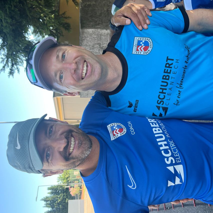 Zwei Männer posieren für ein Foto, beide tragen blaue T-Shirts. Das T-Shirt links trägt Nike, und das T-Shirt rechts trägt Schuber. Im Hintergrund sind Bäume und ein Gebäude zu sehen.