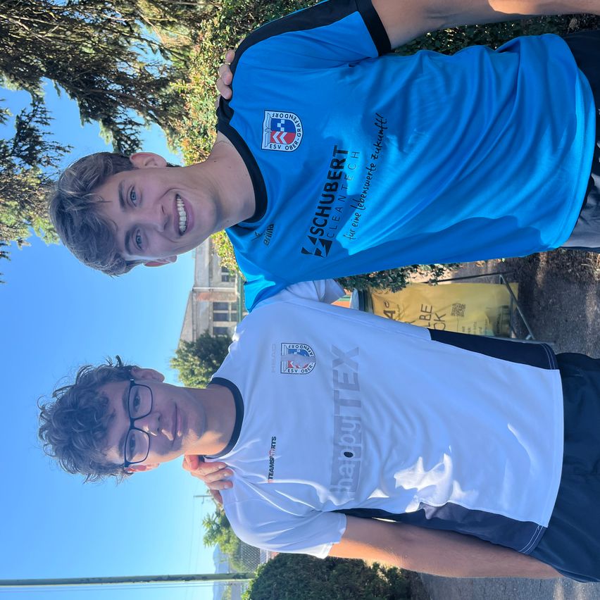 Zwei junge Männer stehen nah beieinander, einer trägt ein blaues Shirt mit 'SCHUBERT CLEANTECH', der andere ein weißes mit 'happyTEX'. Sie haben beide ein Lächeln im Gesicht. Hinter ihnen ist ein Baum und ein Gebäude mit einem gelben Banner.