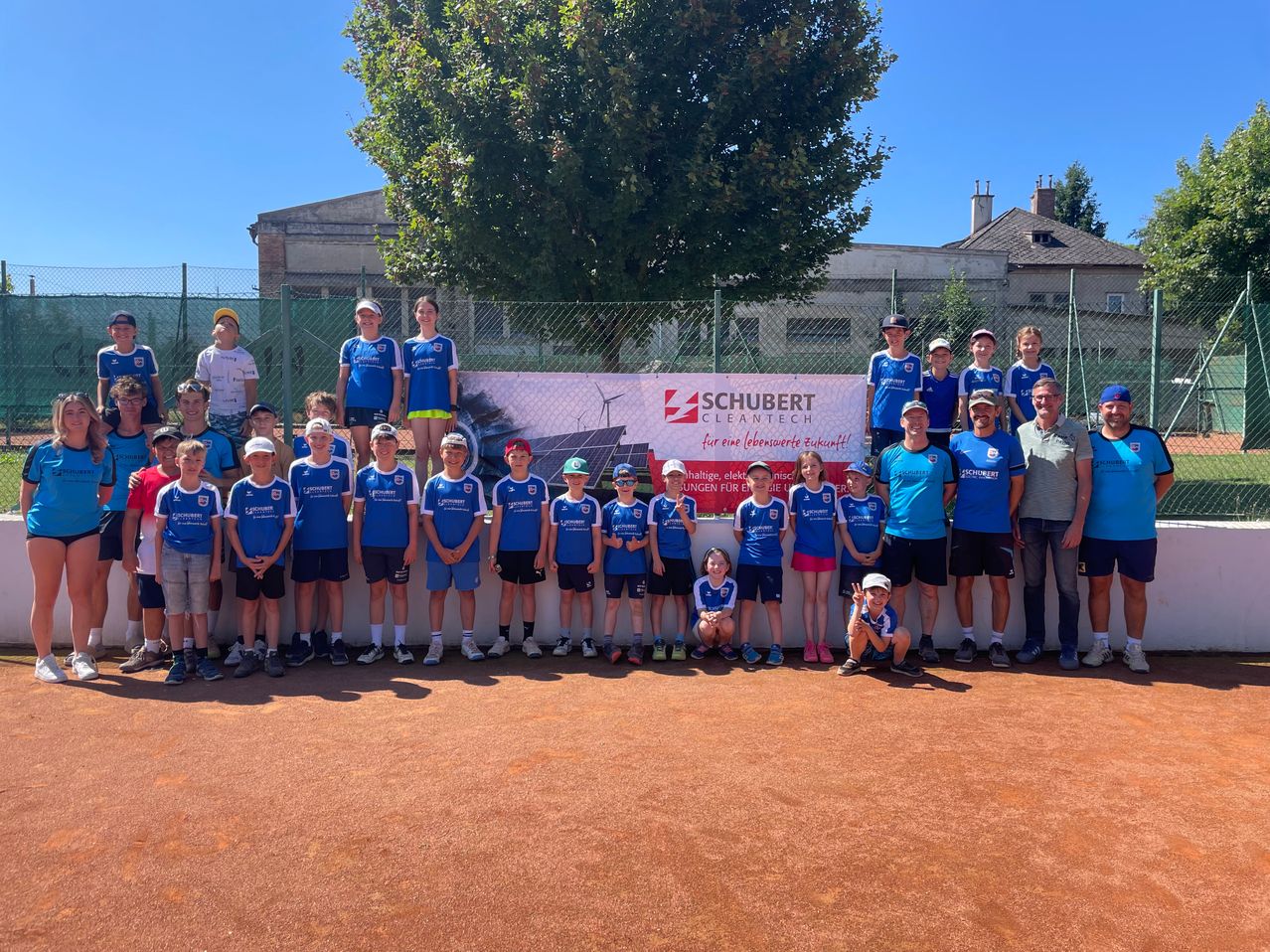 Eine Gruppe junger Athleten in blauen Uniformen posiert für ein Foto auf einem Sportplatz mit einem Banner, das 'SCHUBERT CLEANTECH' besagt. Dahinter umgibt ein Zaun das Gelände mit Gebäuden und Bäumen im Hintergrund.