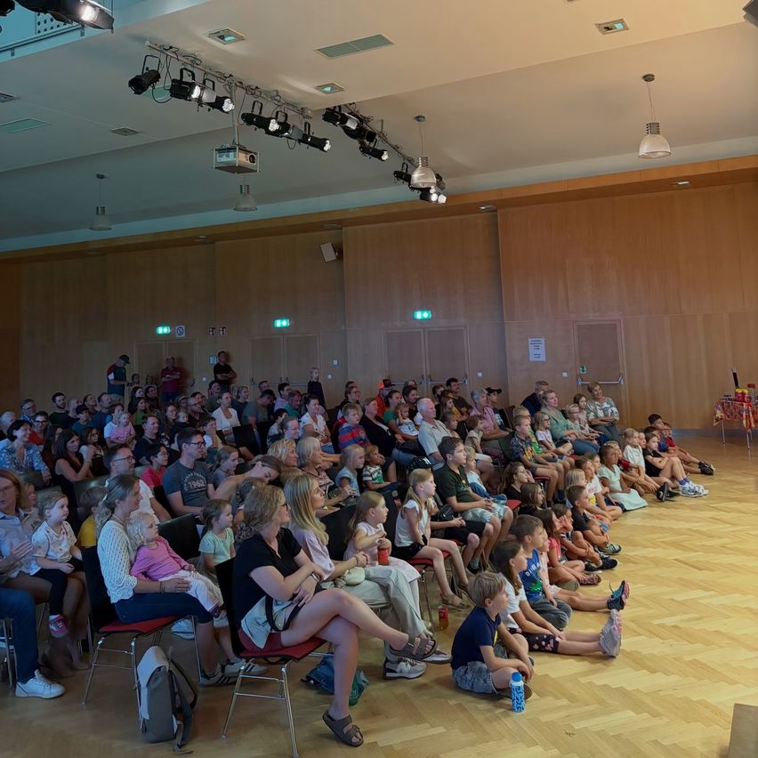 Eine große Gruppe von Menschen, einschließlich Kindern, sitzt in Reihen in einem gut beleuchteten Auditorium und sieht sich eine Präsentation an. Der Raum hat Holzwände und einen Holzboden.