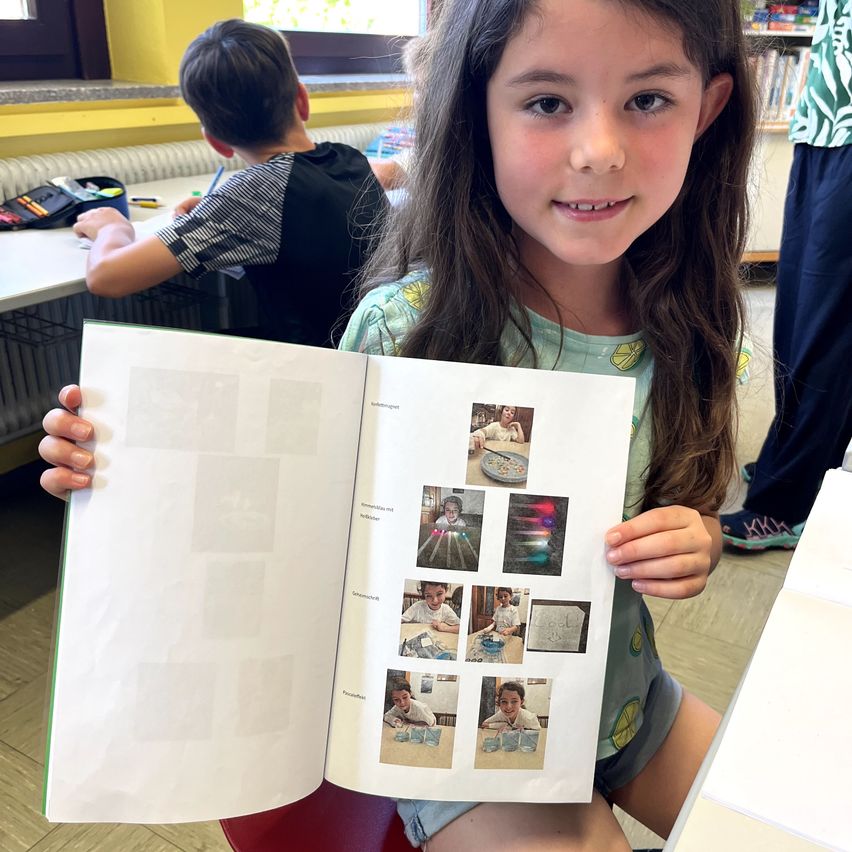 Ein junges Mädchen sitzt in einem Klassenzimmer, lächelt und hält ein offenes Buch mit Bildern und Zeichnungen in den Händen. Hinter ihr sitzt ein Junge am Schreibtisch.