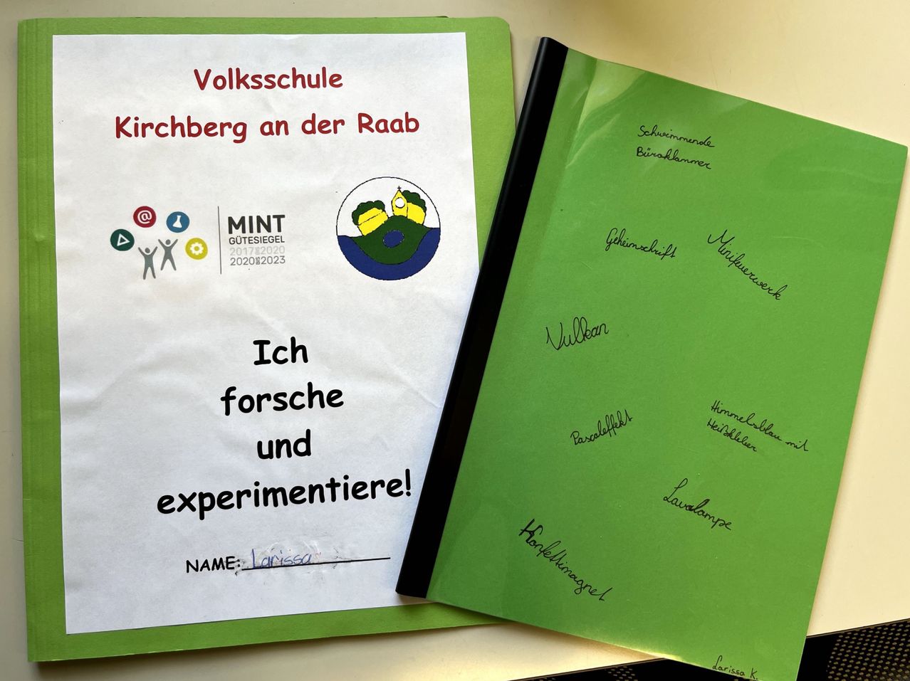 Ein weißes Papier mit grünem Rand steht 'Volksschule Kirchberg an der Raab'. Darunter befindet sich ein Logo und Symbole. Ein grüner Ordner ist geöffnet, mit handgeschriebenen Notizen und Unterschriften.