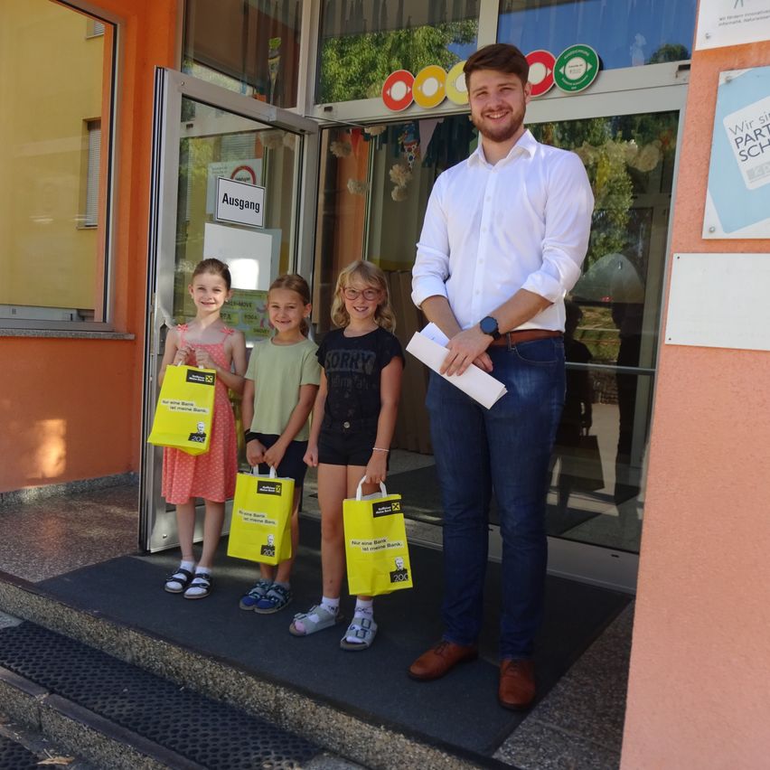 Ein Mann steht mit drei Mädchen vor einem Gebäude und lächelt. Sie alle halten gelbe Taschen. Das Gebäude hat eine Glastür mit Schildern darüber.