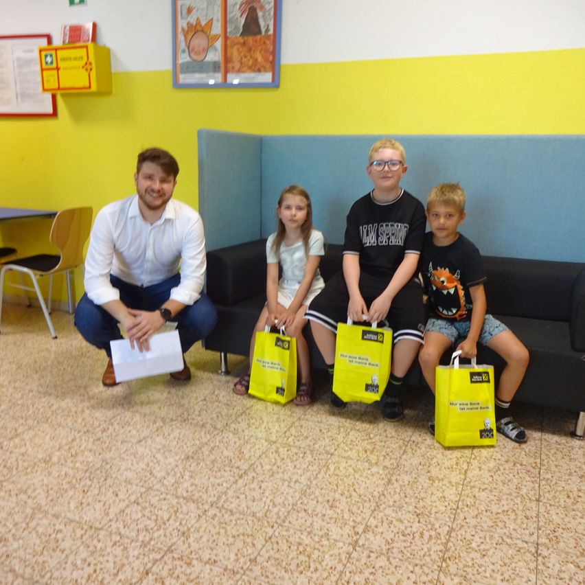 Ein Mann und drei Kinder sitzen auf einer Couch und halten gelbe Taschen. Sie alle scheinen zu lächeln und posieren für ein Foto. Der Raum hat eine gelb-blaue Wand und einen Schreibtisch mit einem Stuhl im Hintergrund.
