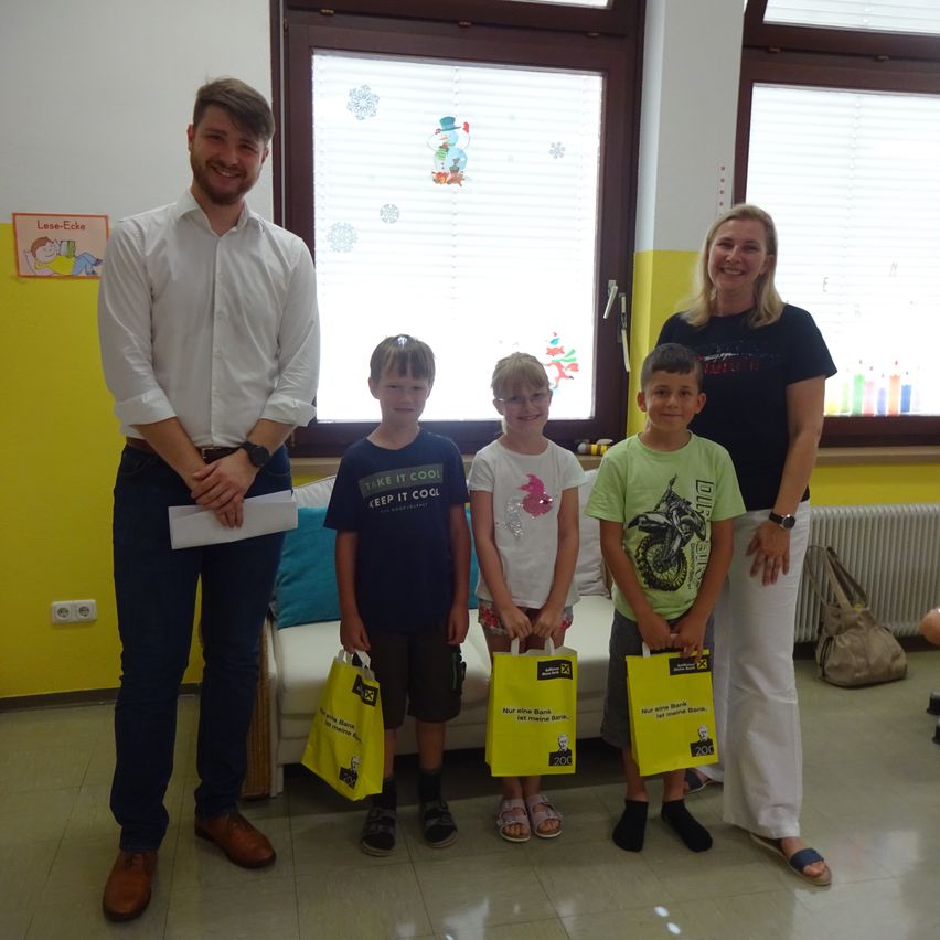 Eine Gruppe von Kindern und Erwachsenen steht lächelnd in einem Raum. Ein Mann hält ein Papier, und jedes Kind hält einen gelben Beutel. Hinter ihnen zeigt ein Fenster Schneeflocken.